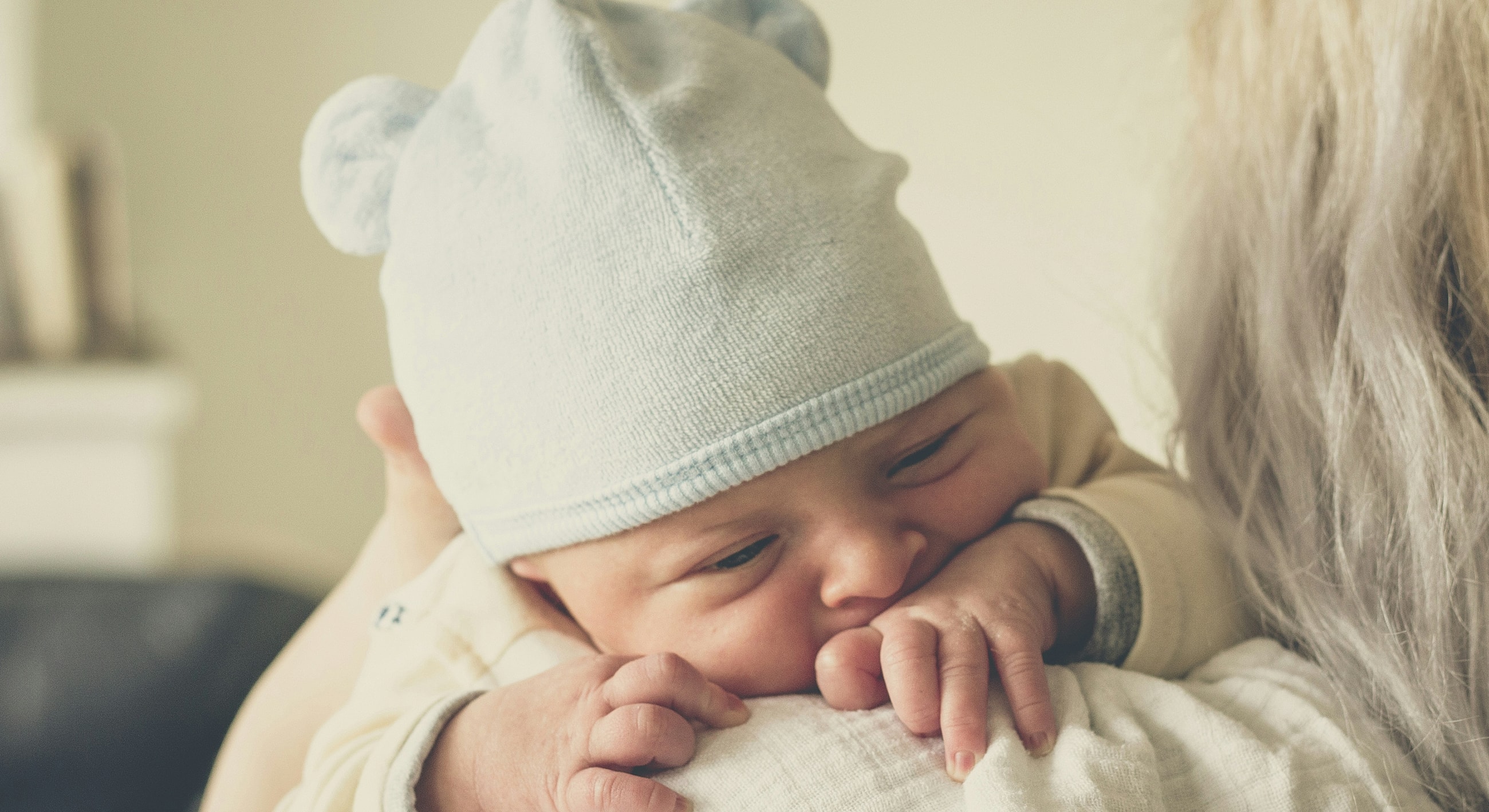 Baby with blue hat resting on caregiver's shoulder.