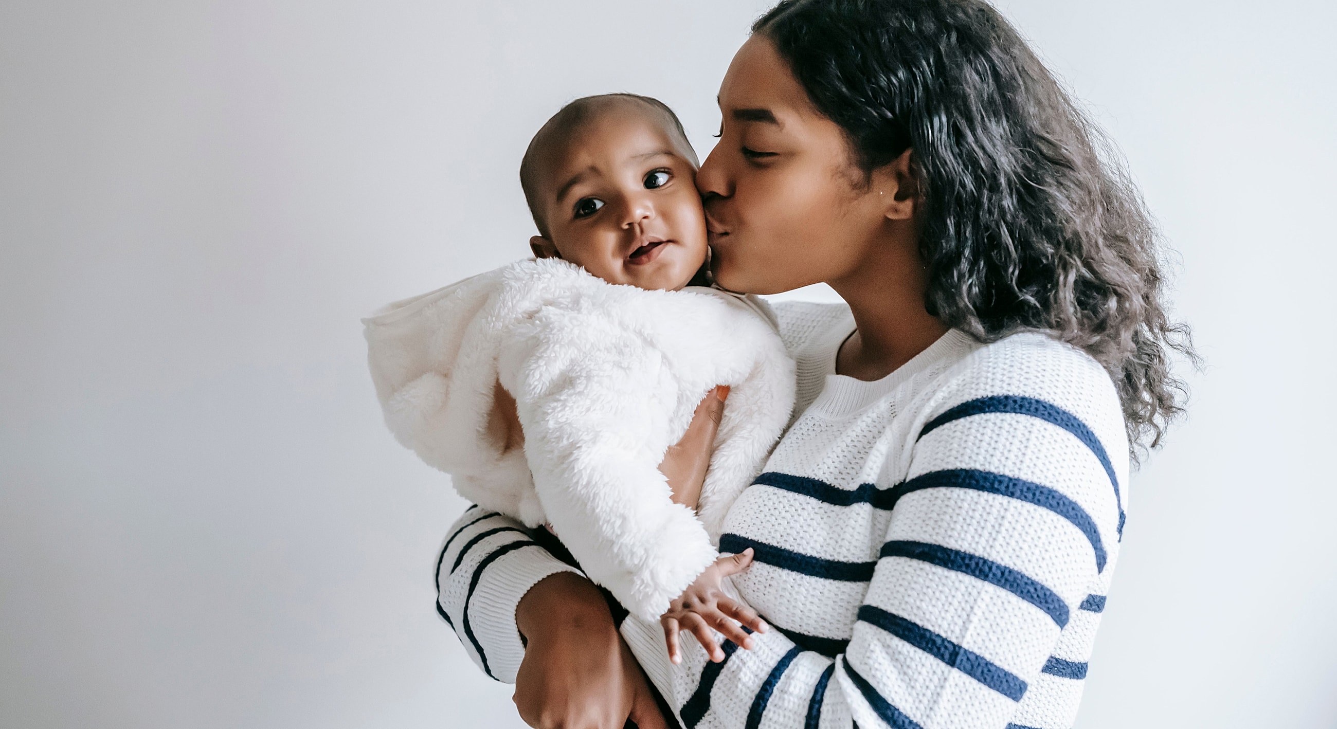 Mother kissing baby in cozy white outfit.